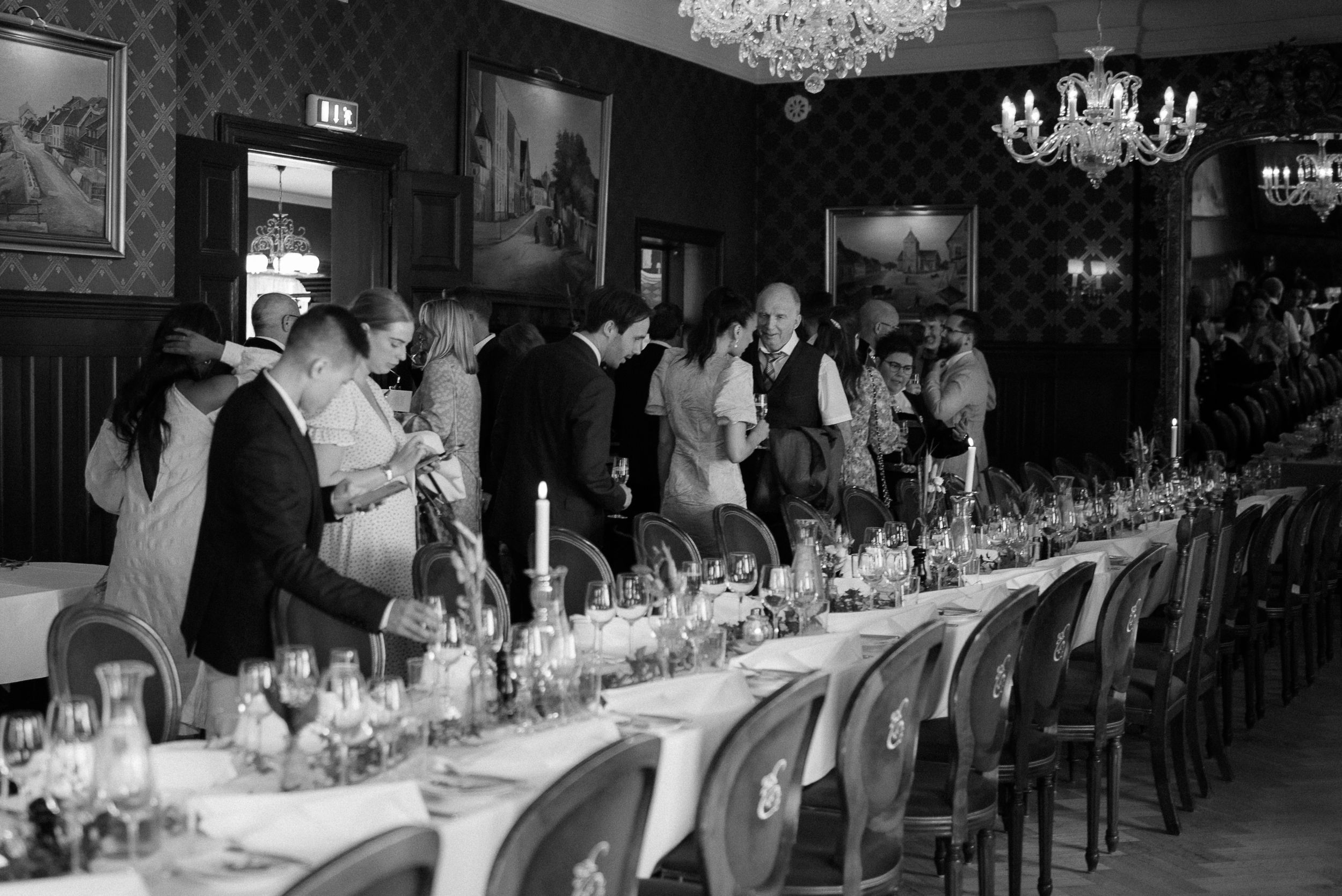 Black and white photo of wedding guests mingling around an elegantly set long dining table.
