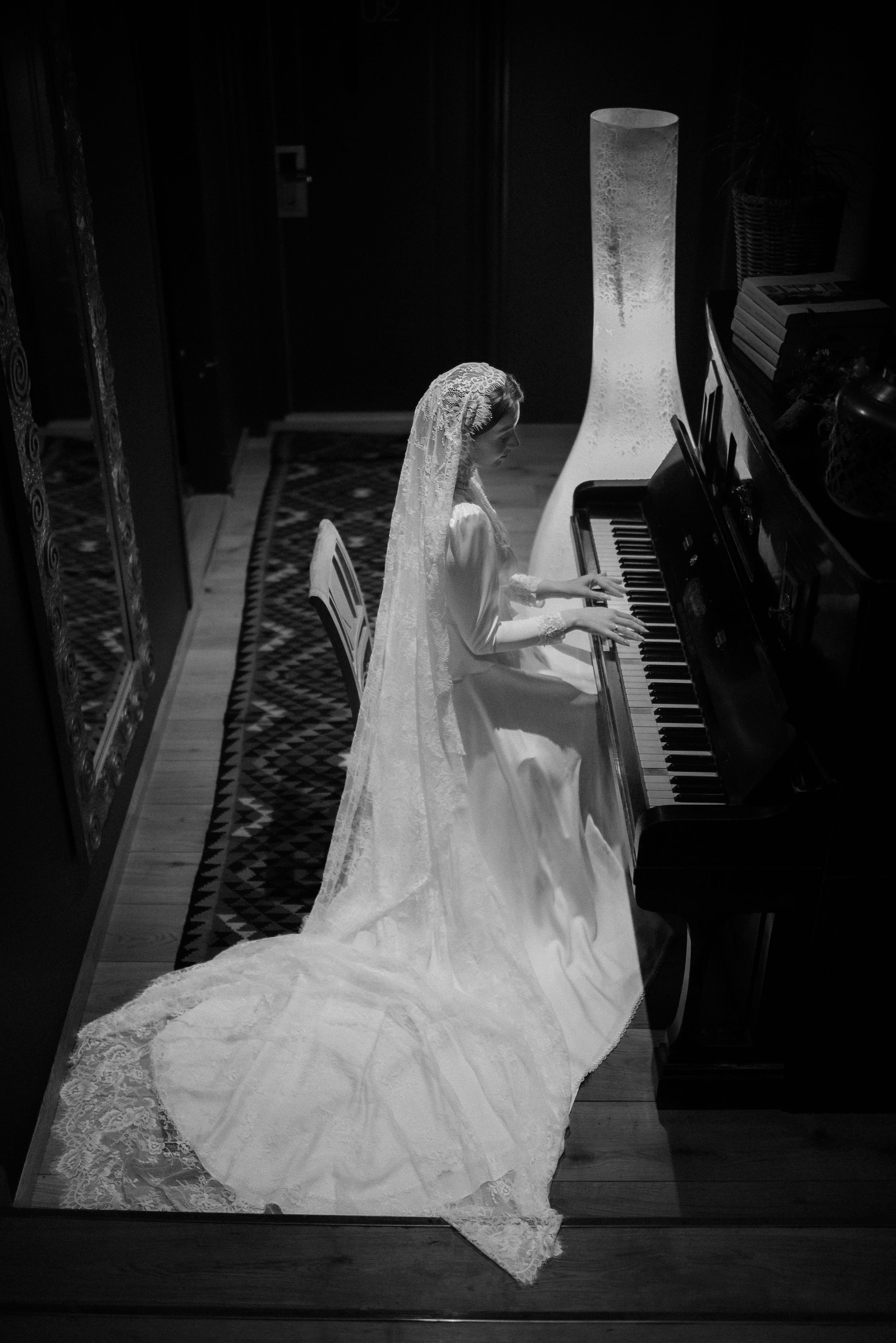 Elegant bride in flowing lace gown playing piano in dramatic black and white photography.