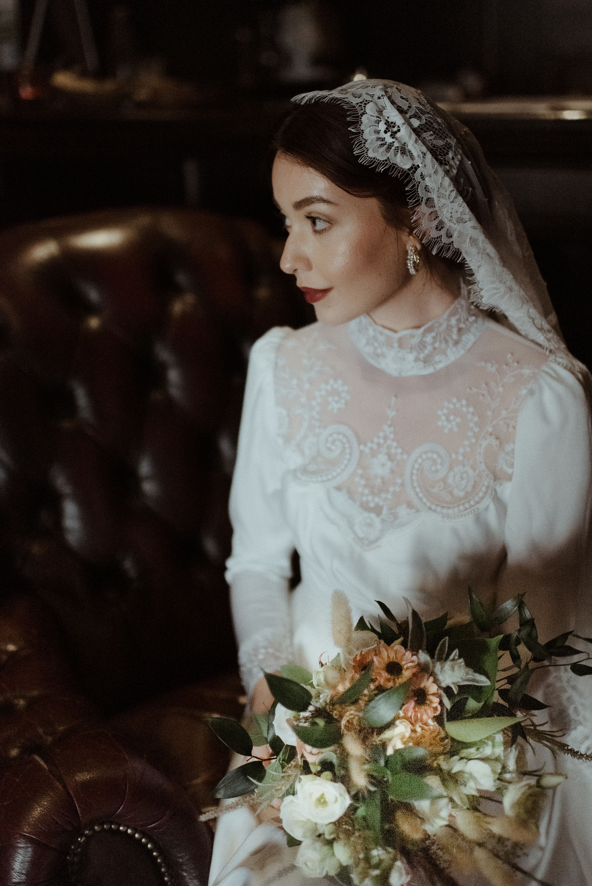 Bride in vintage-style lace wedding dress with bouquet against leather chair.