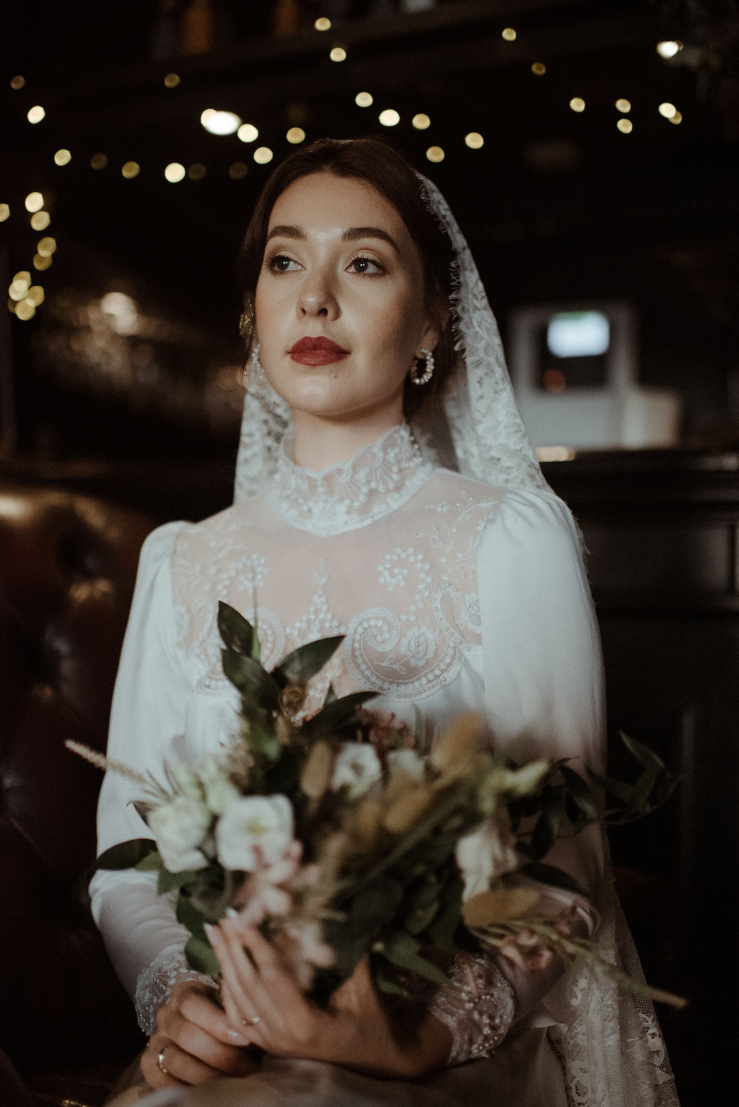 Elegant bride in vintage-inspired lace dress holding floral bouquet with bokeh lights.