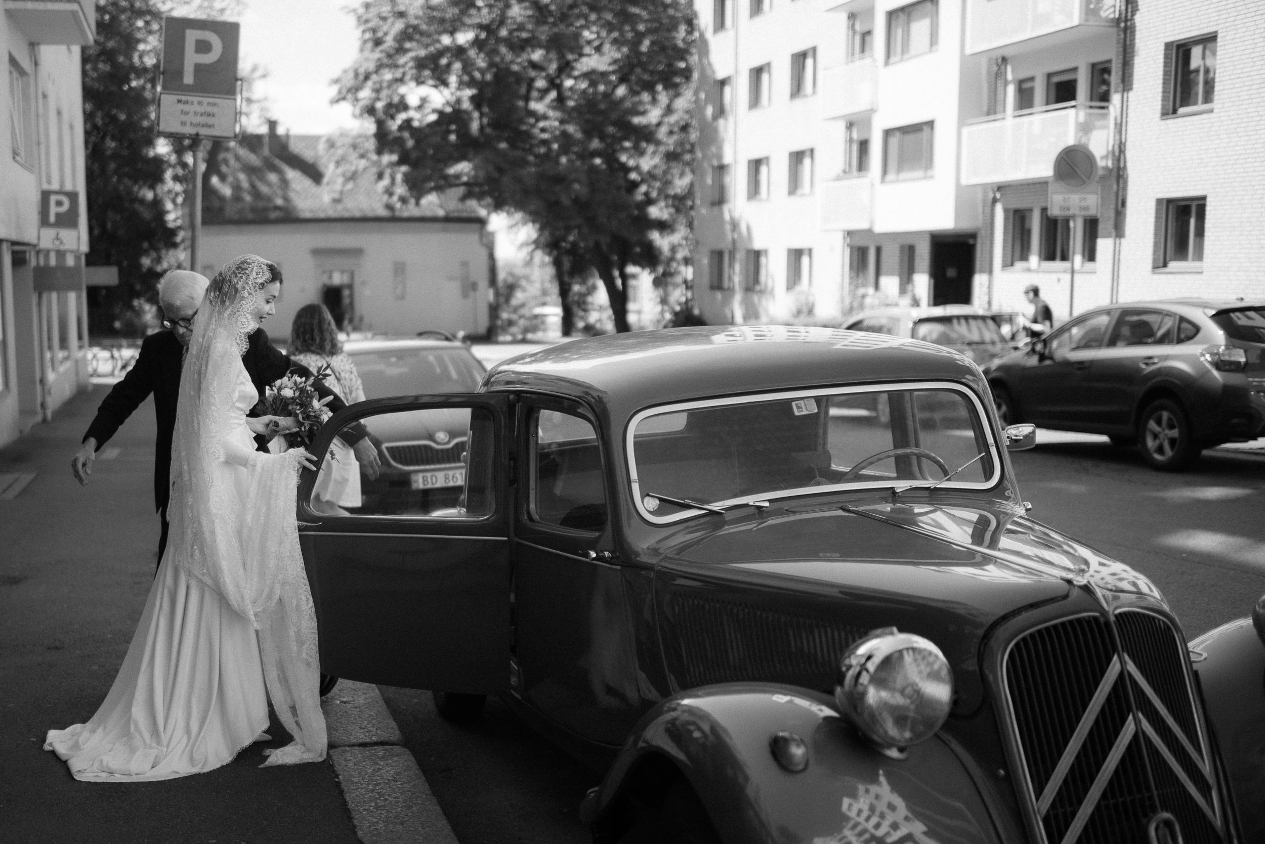 Bride in flowing white dress entering classic Citroen car on city street.
