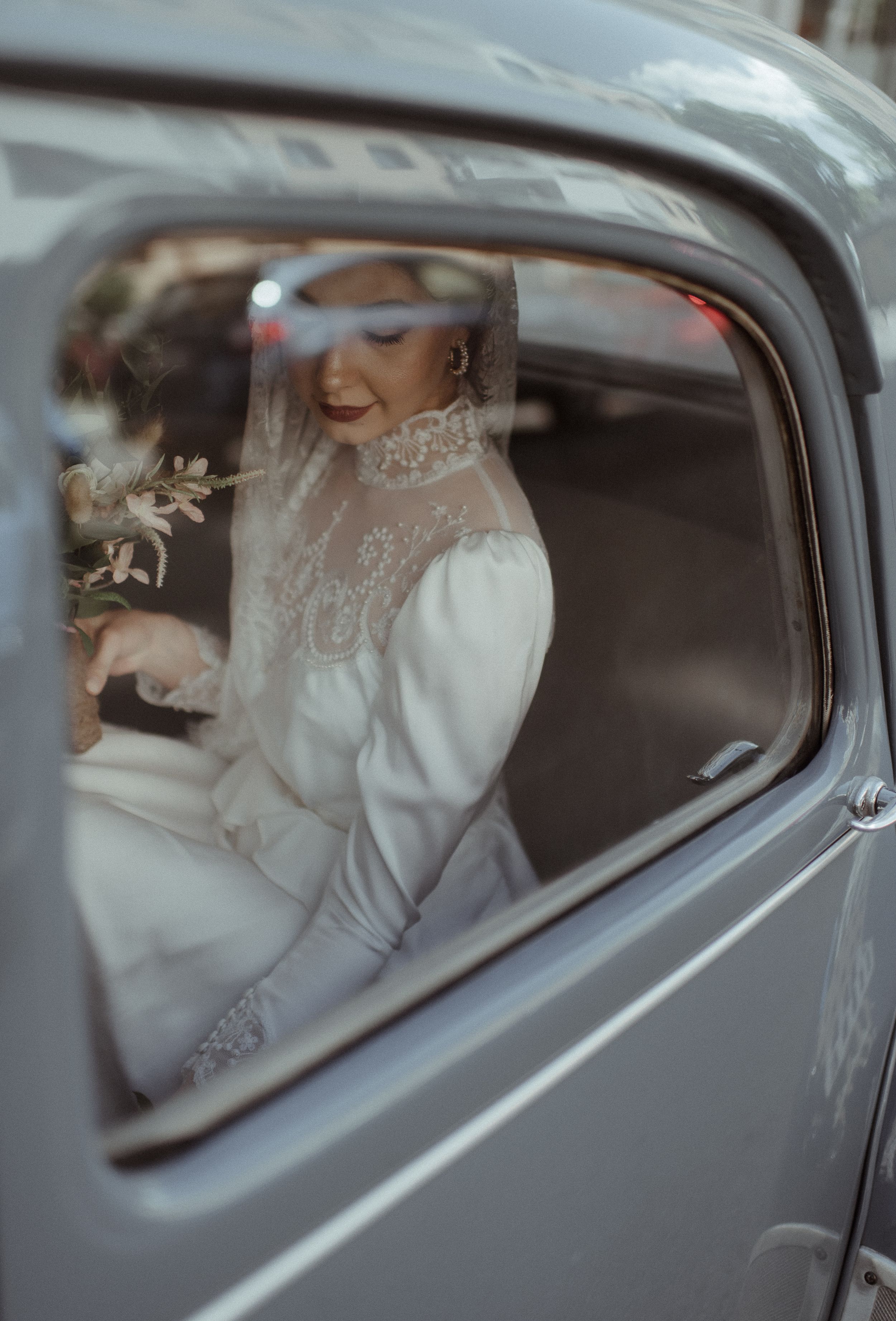 A vintage car window frames a bride in an elegant lace and long-sleeved wedding dress.