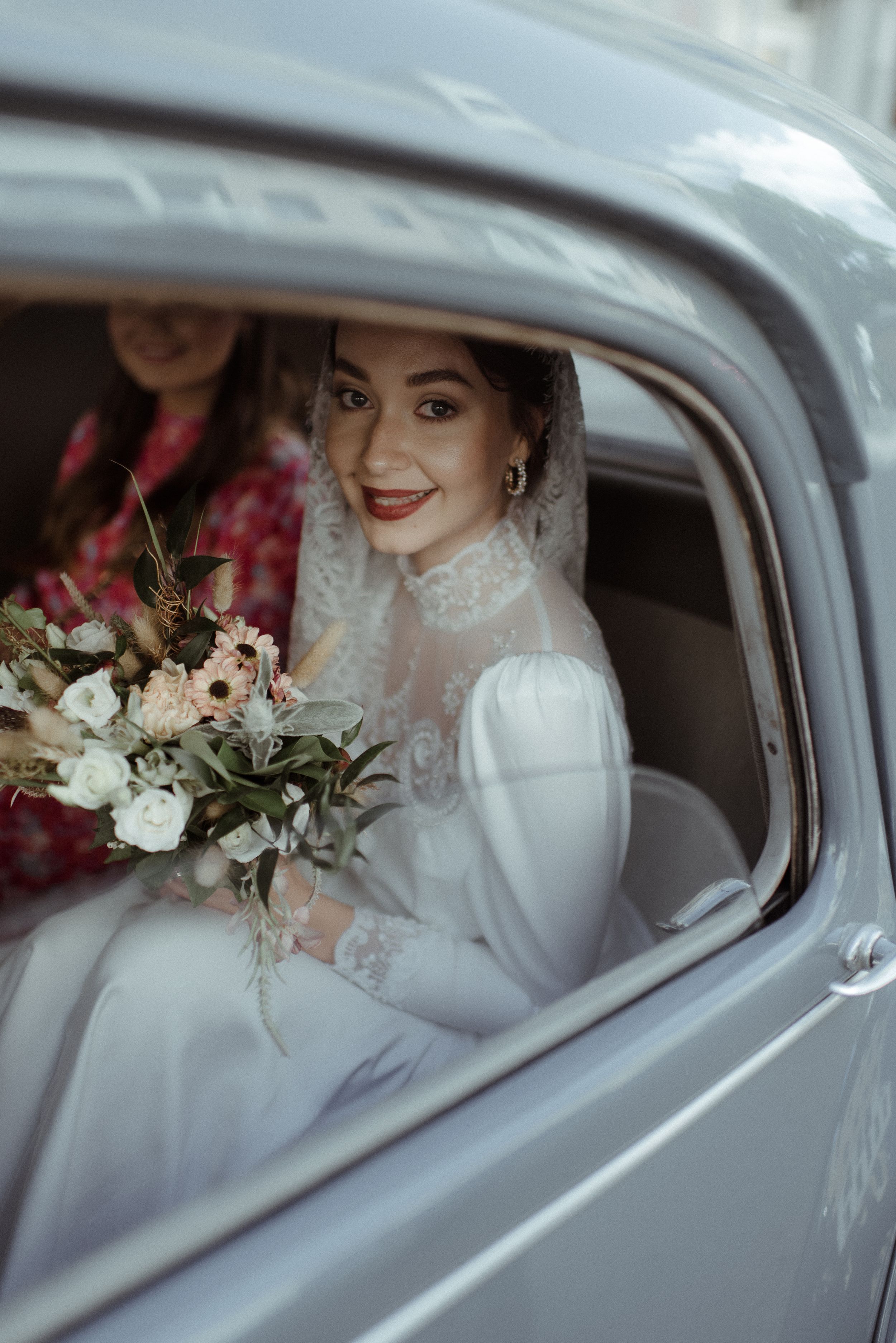 A bride in a classic white gown holds a delicate bouquet while sitting in a vintage car.