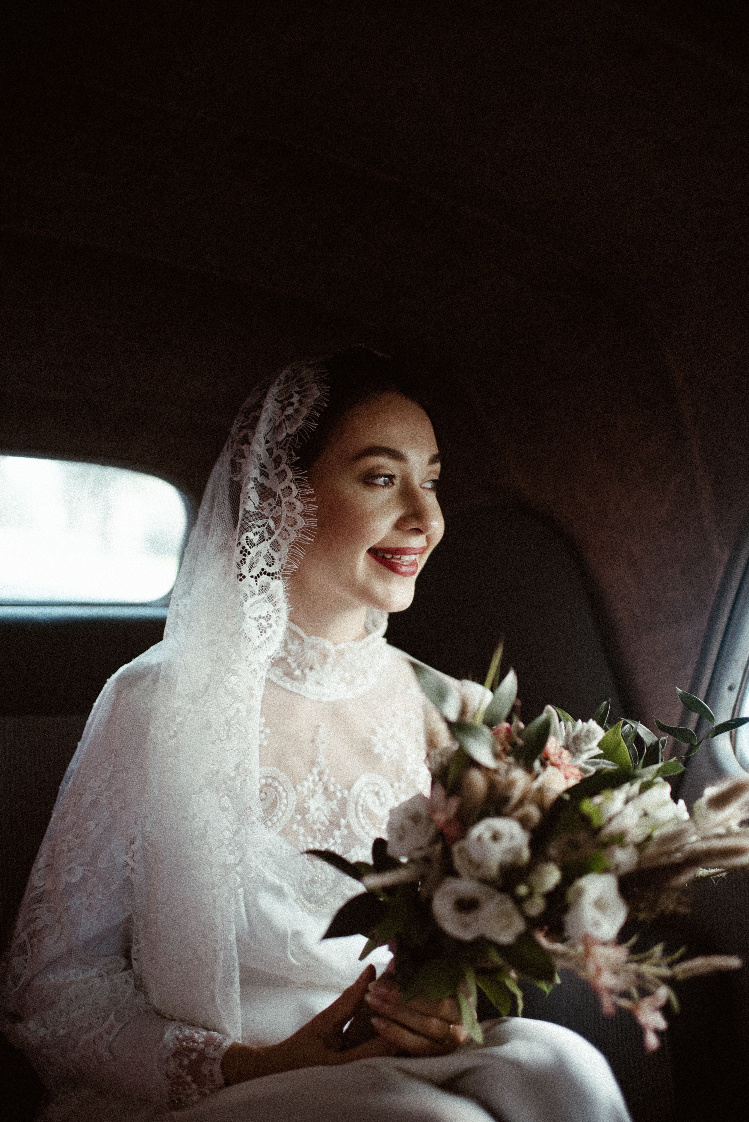 An elegant bride with a lace veil holds a romantic bouquet of flowers in a classic car.