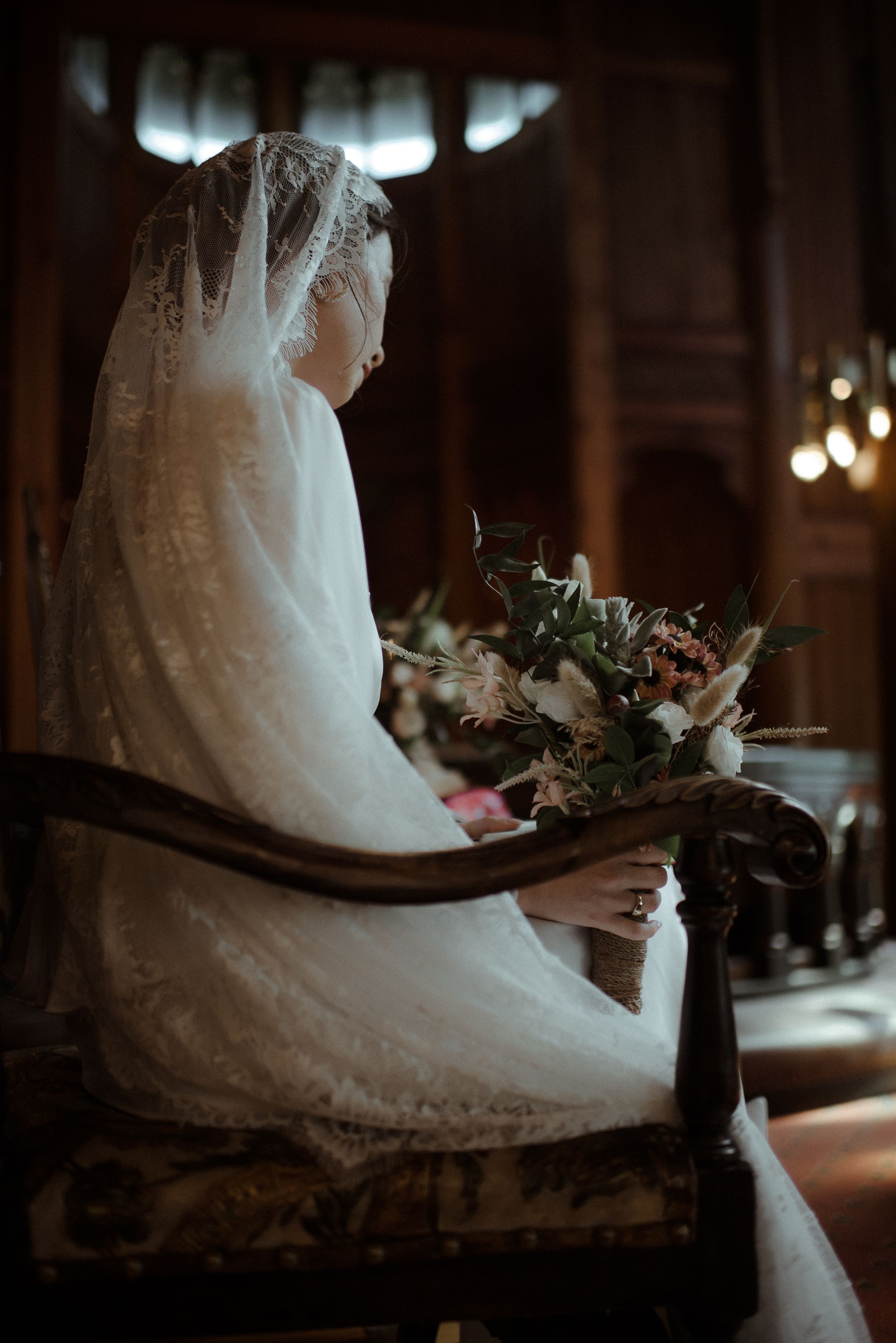 A contemplative side view of a bride in white lace holding her wedding bouquet while seated.
