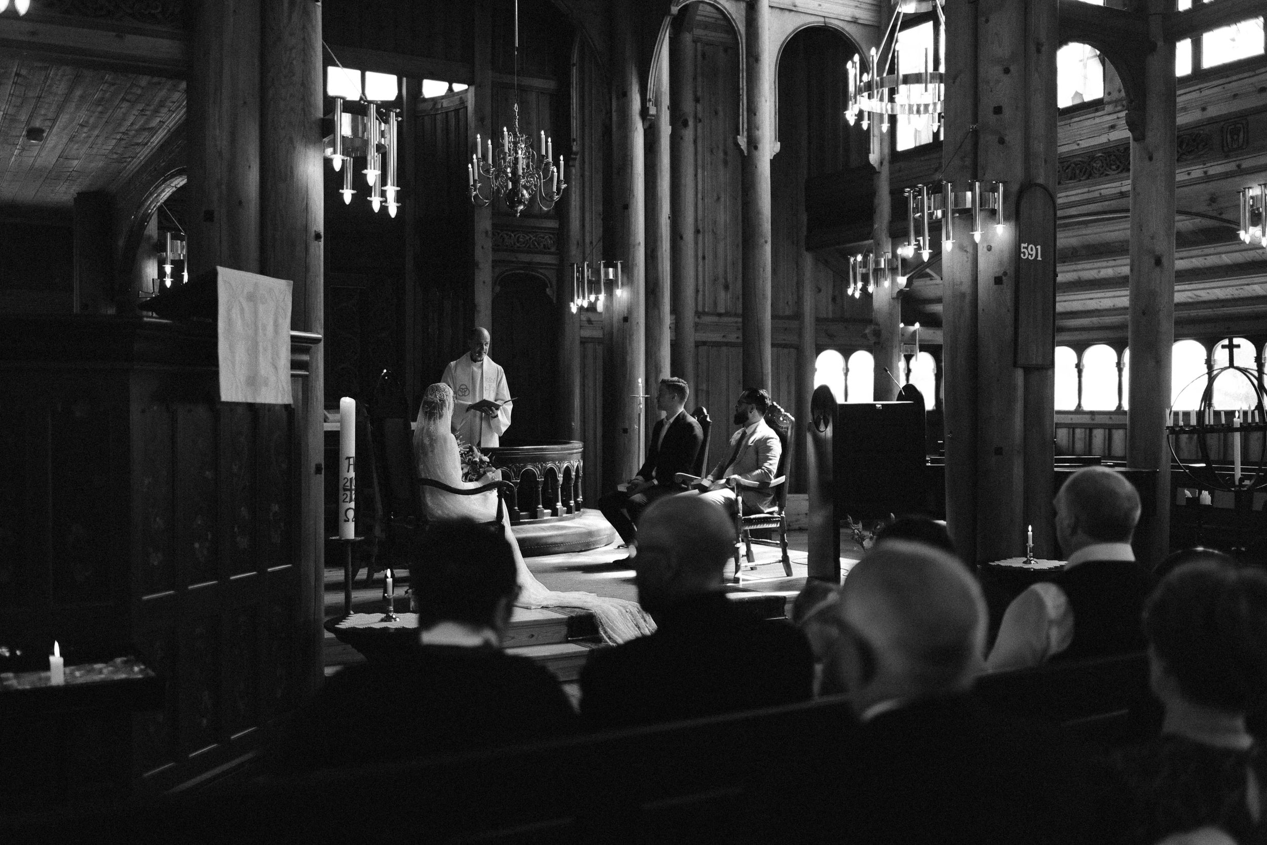 A moody black and white photo captures an interior church ceremony with candlelit fixtures and wooden pews.