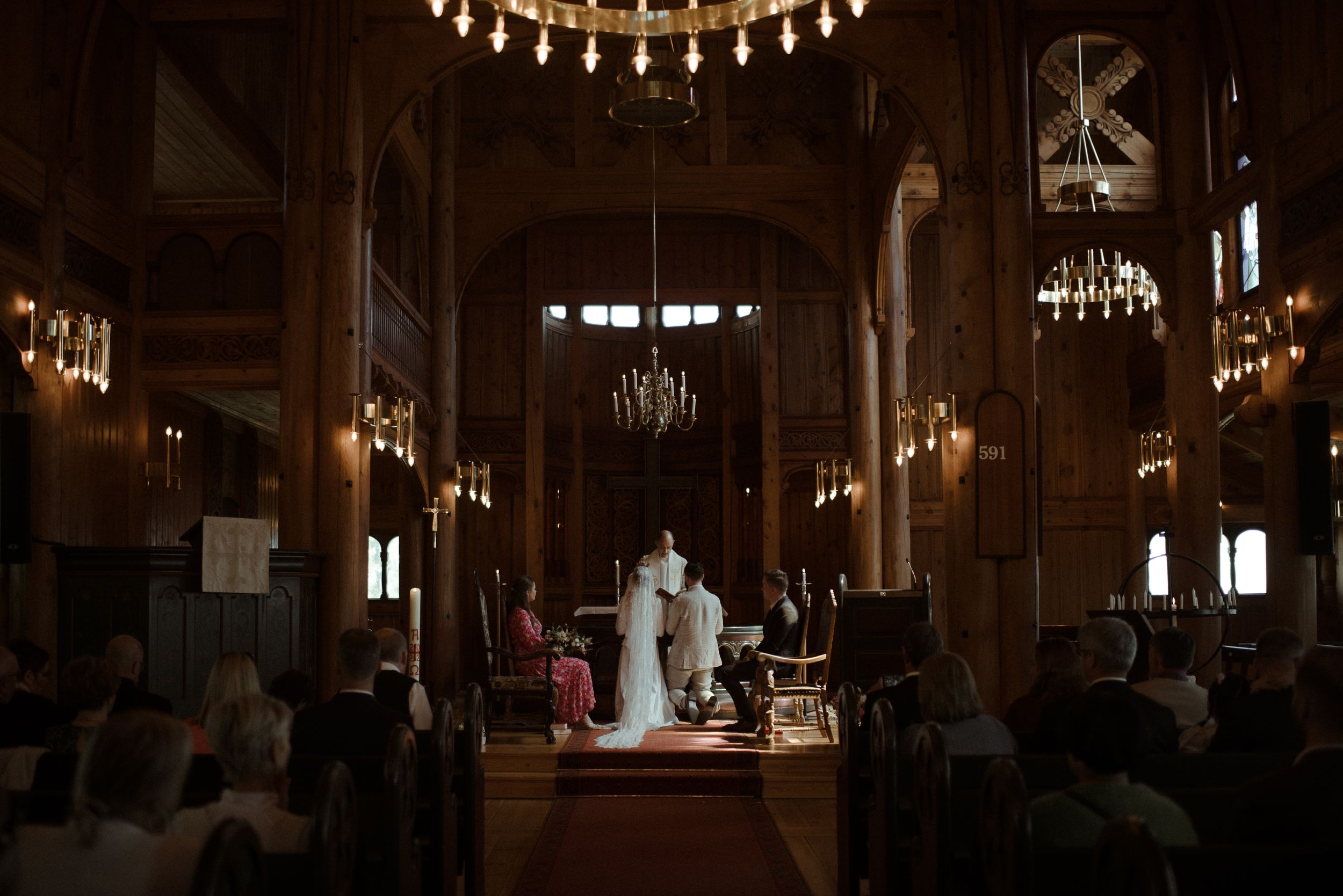 A wedding ceremony viewed from the back of a chapel with elegant chandeliers and wooden details.