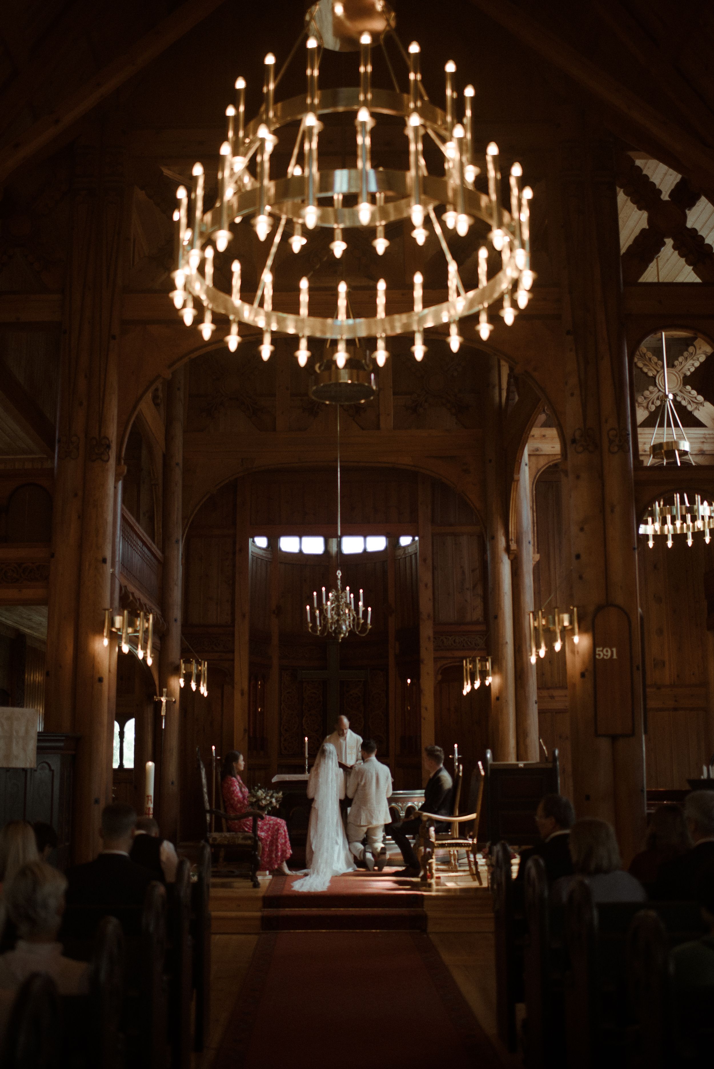 A grand chandelier illuminates a church aisle during a wedding ceremony under dramatic wooden arches.
