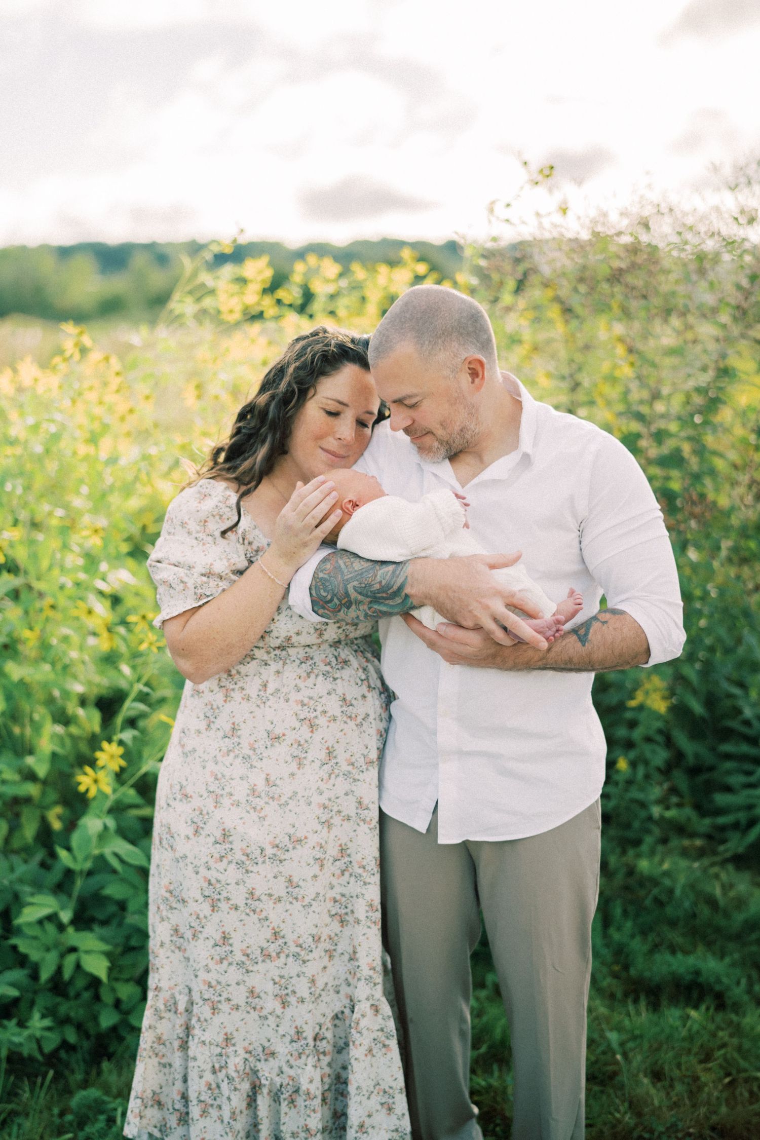 A couple holds their newborn baby while standing in a field of yellow wildflowers on a sunny summer day.