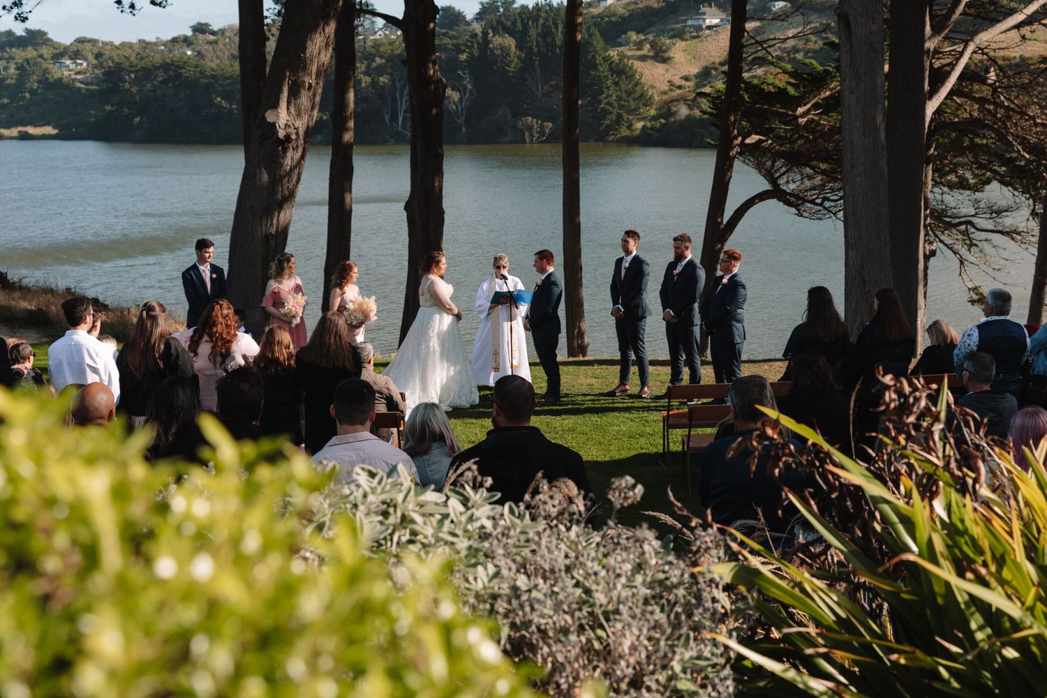 Outdoor wedding ceremony by a lake with guests seated among tall trees.