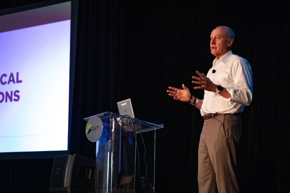A speaker delivers a presentation at a podium on stage with a projection screen display in the background.