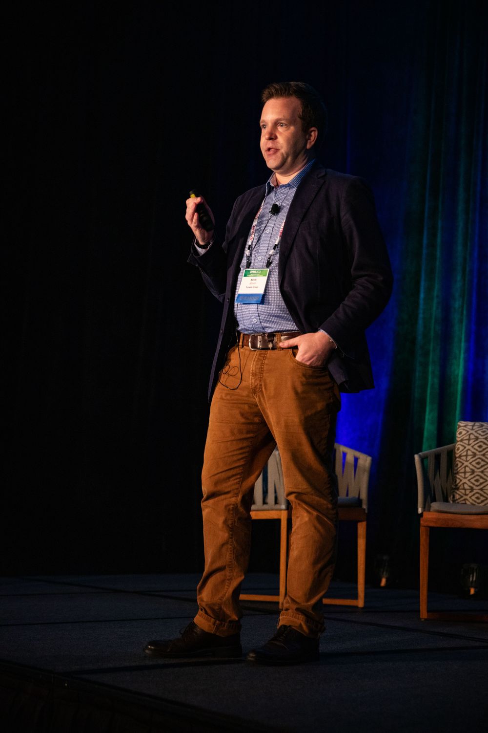 Speaker on stage at business conference wearing navy blazer and brown pants gestures while giving presentation.