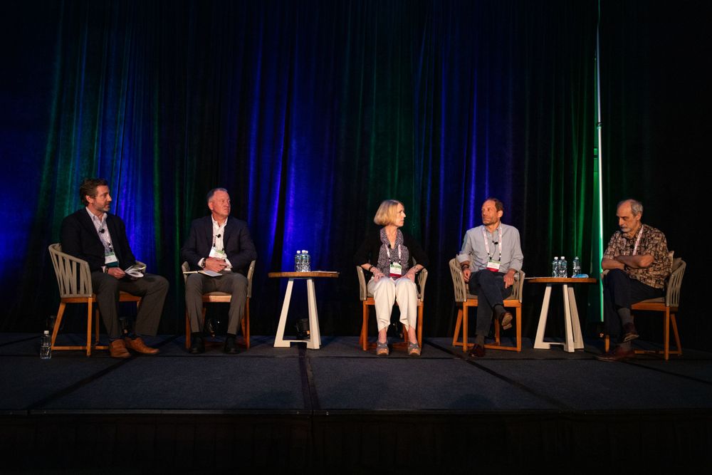 Five speakers seated on stage during a panel discussion with blue and green lighting effects.