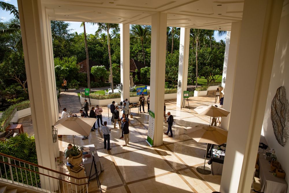 People gather on an elegant columned terrace overlooking manicured gardens and palm trees at a luxury venue.