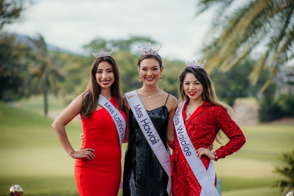 Three pageant contestants pose together outdoors wearing formal dresses in red and black against a tropical background.