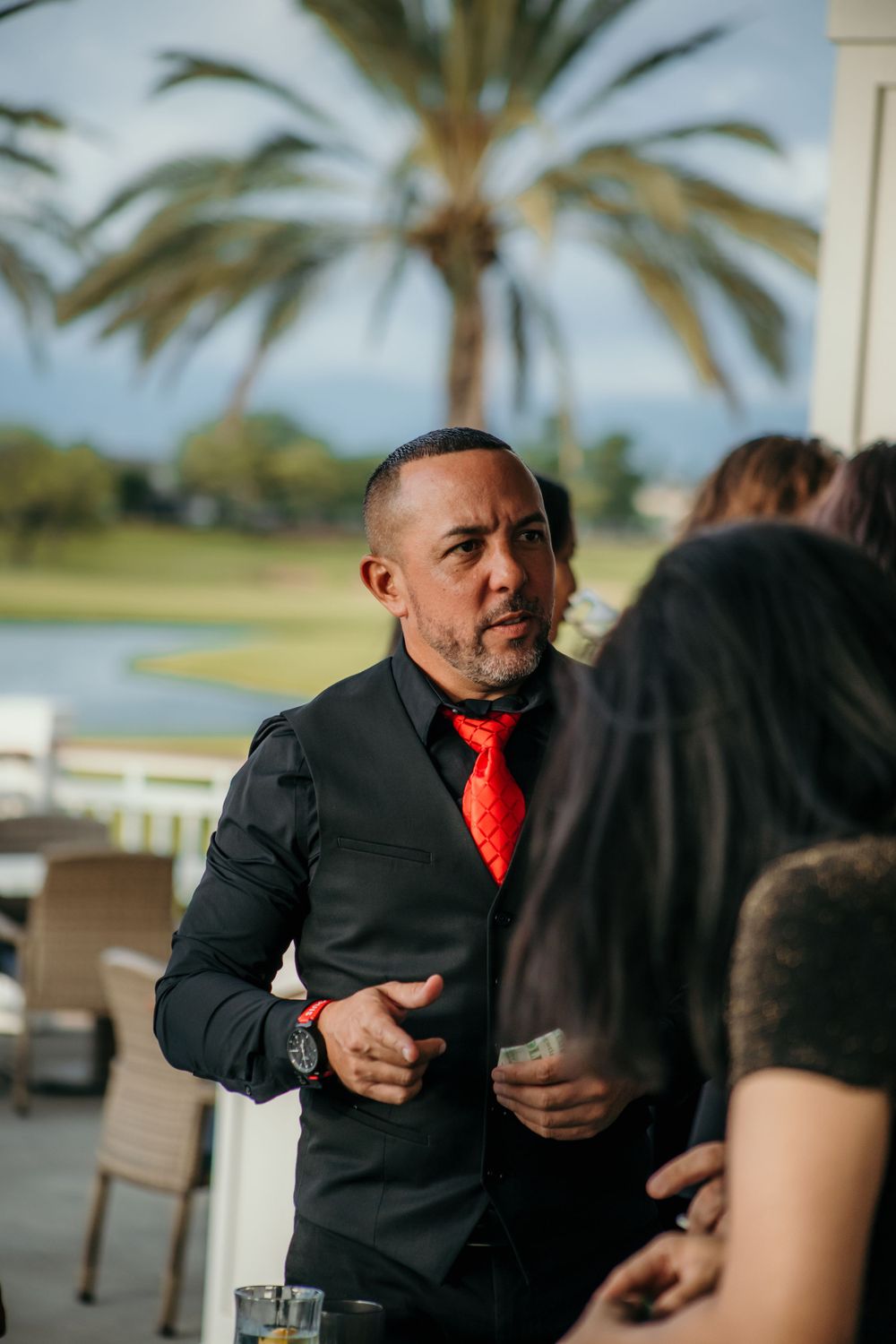 A person in a black suit and red tie gestures while engaged in conversation outdoors with palm trees in the background.