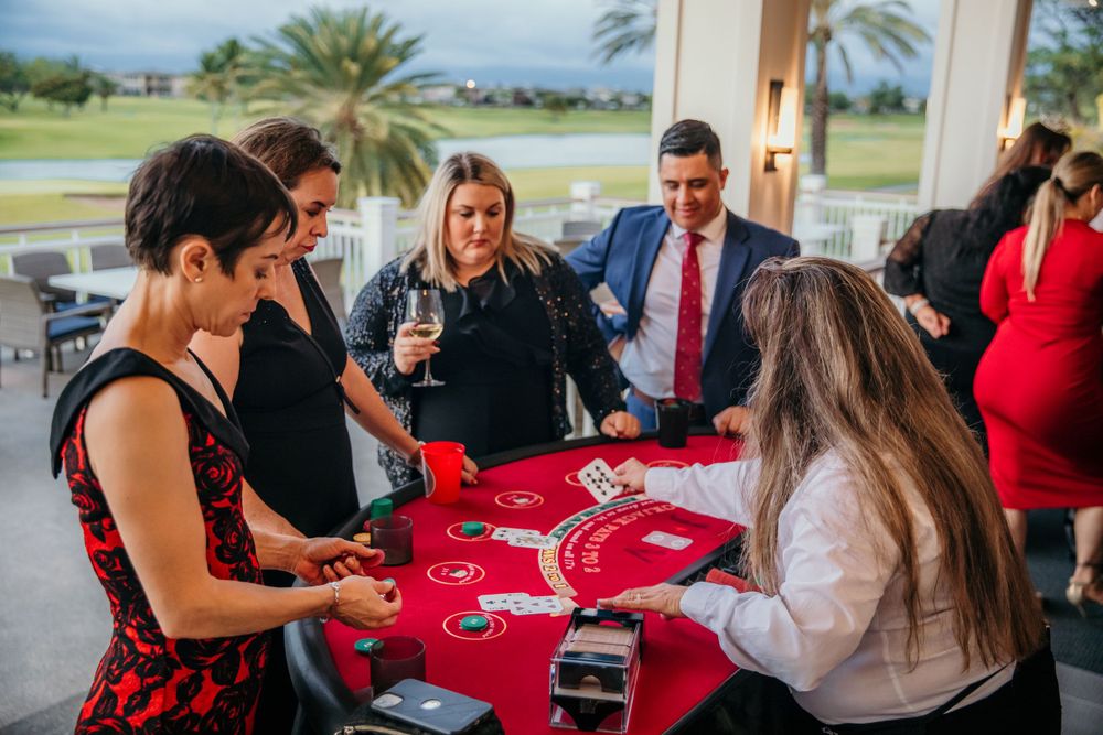 People gathered around a red casino table enjoying drinks and card games with palm trees visible through windows in background.