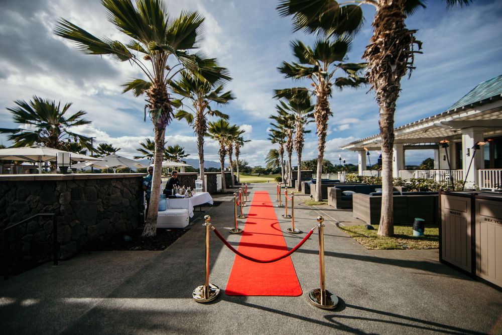A red carpet path between palm trees leads to an oceanfront venue with stanchions and ropes along a sunny boardwalk.