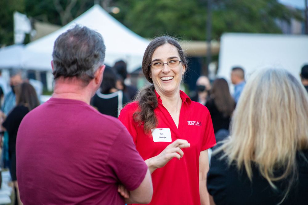 A person in a red polo shirt engages in conversation at an outdoor event with white tents in the background.
