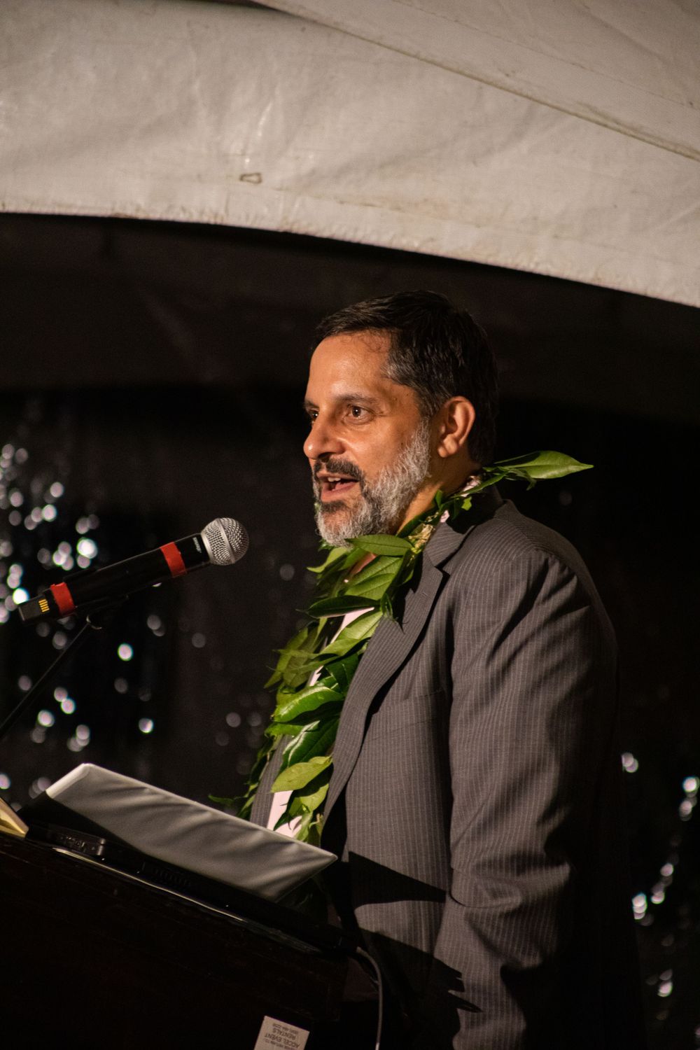 A speaker in a grey suit with traditional lei addresses audience from podium at evening event.