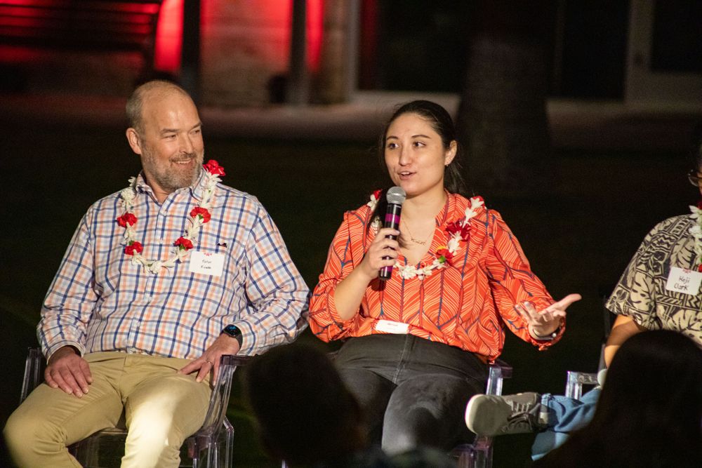 Two speakers share a stage during an evening presentation wearing leis and using microphones.