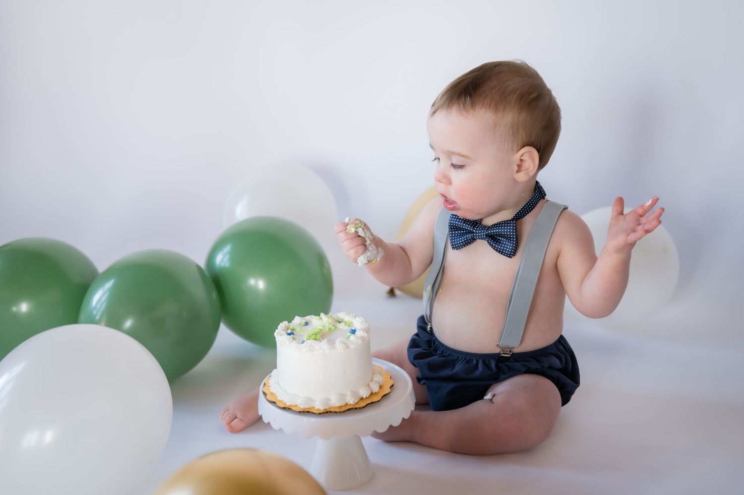 Baby in navy suspenders and bow tie sits with cake and green and white balloons for a special cake smash photo session.