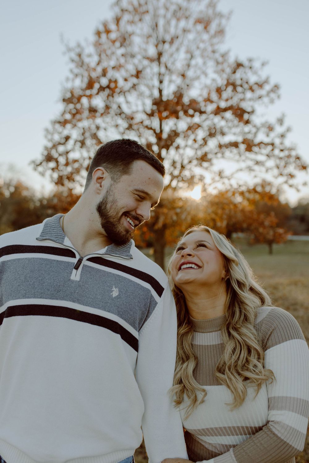 A couple shares a joyful laugh together during an autumn photoshoot with golden trees in the background.