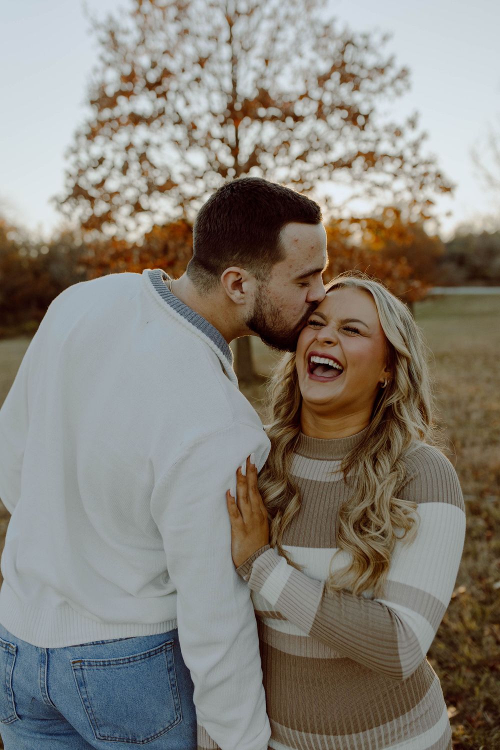A couple shares a joyful moment laughing together outdoors during autumn, wearing cozy sweaters against a backdrop of fall trees.