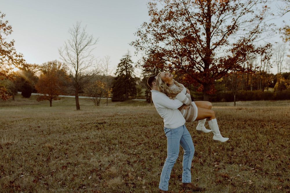 Two people share a joyful moment in a golden-lit field at sunset with autumn trees in the background.