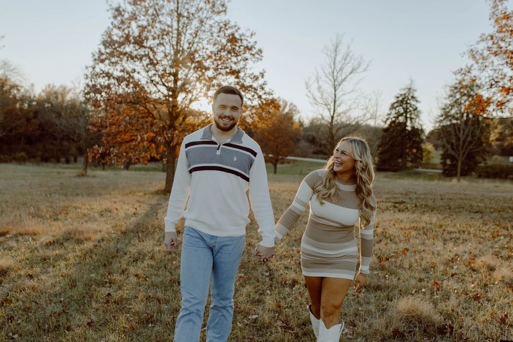 A couple in coordinating sweaters walks hand in hand through an autumn field with golden trees in the background.