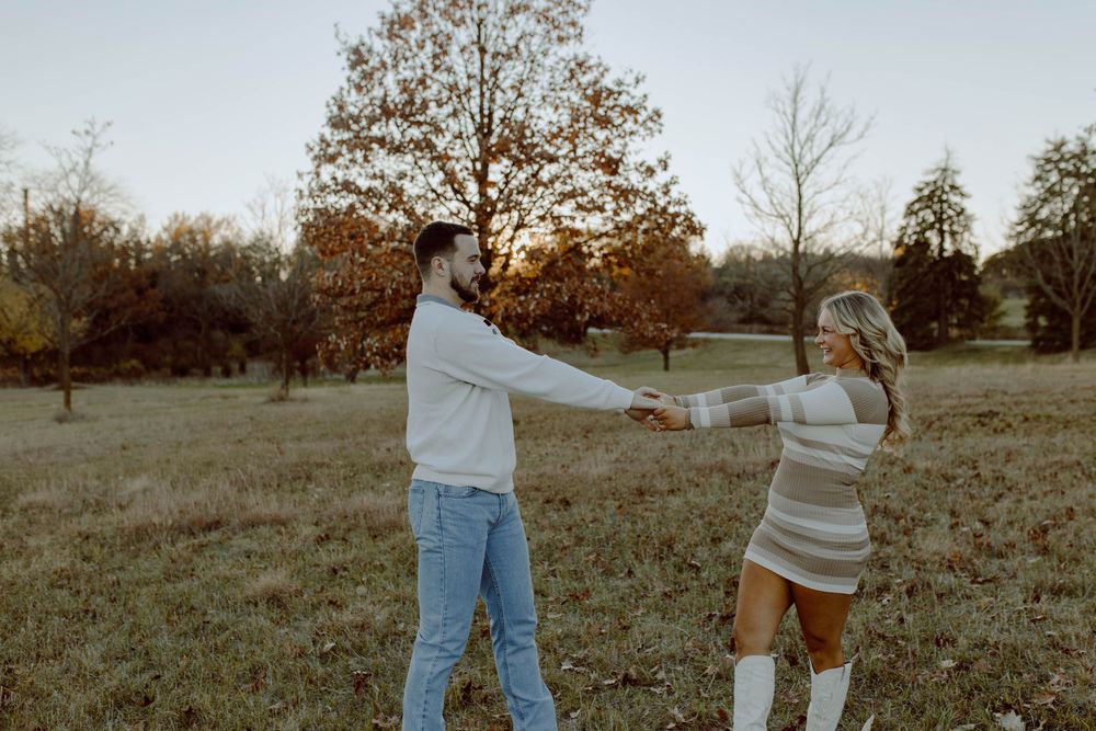Two people hold hands and dance together in an autumn field during golden hour with trees in the background.