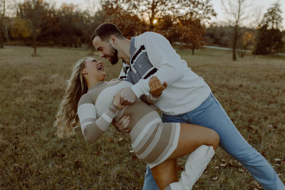 A couple wearing casual outfits embraces playfully in a fall field during golden hour.
