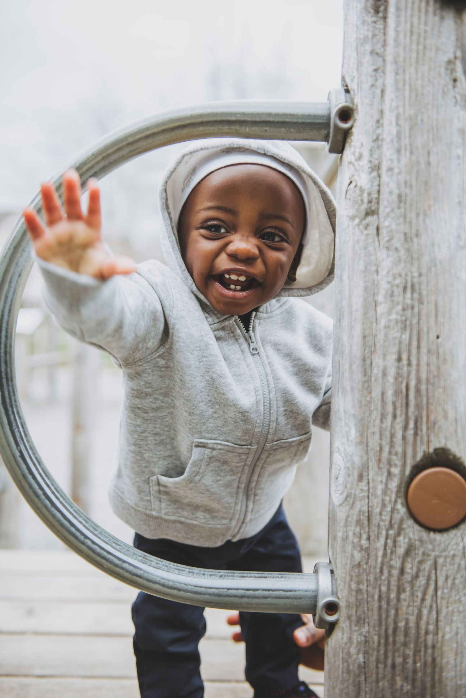A happy toddler in a hoodie smiles and waves enthusiastically while playing on a wooden structure at the playground.
