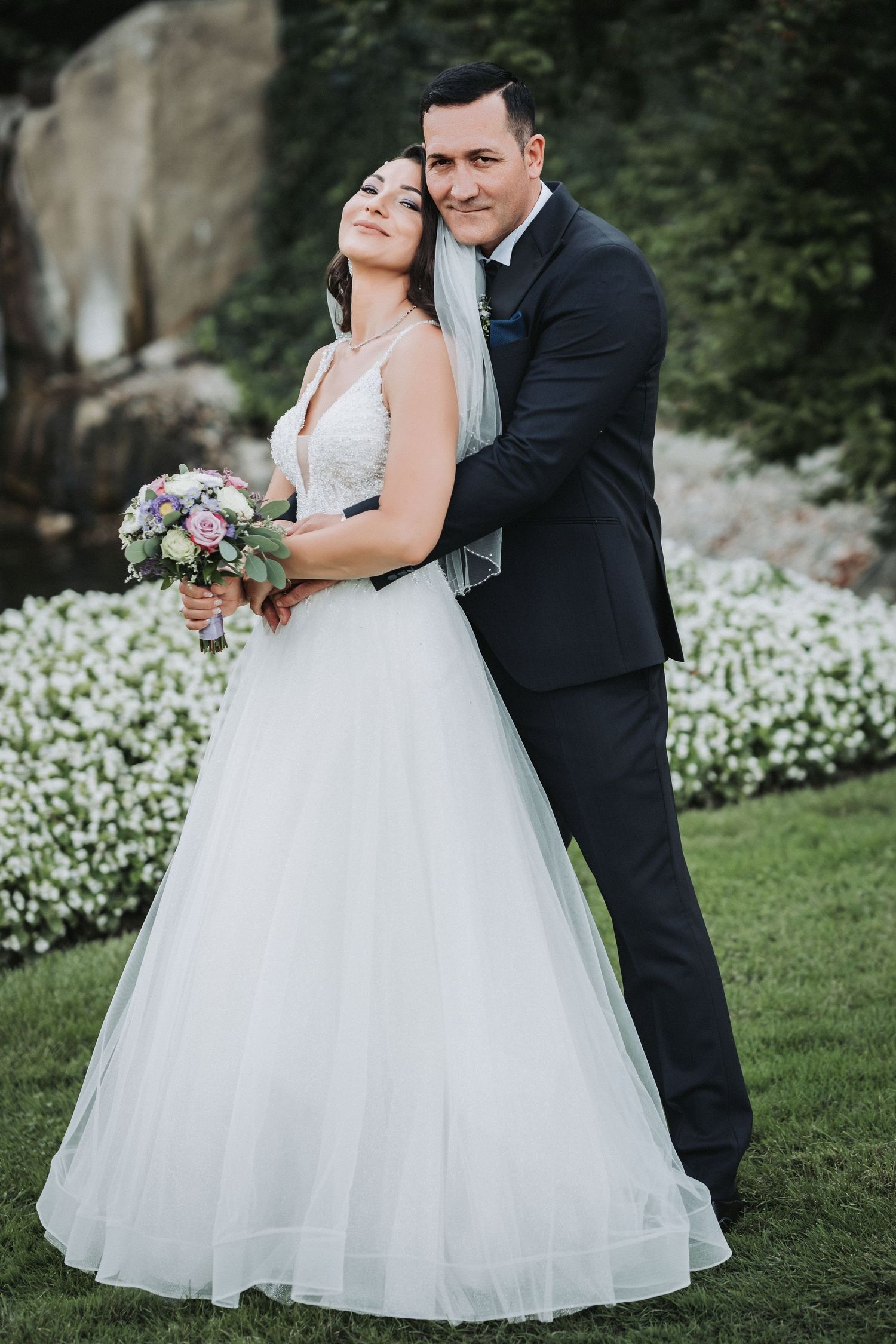 Romantic wedding portrait of couple embracing in garden with white floral backdrop.