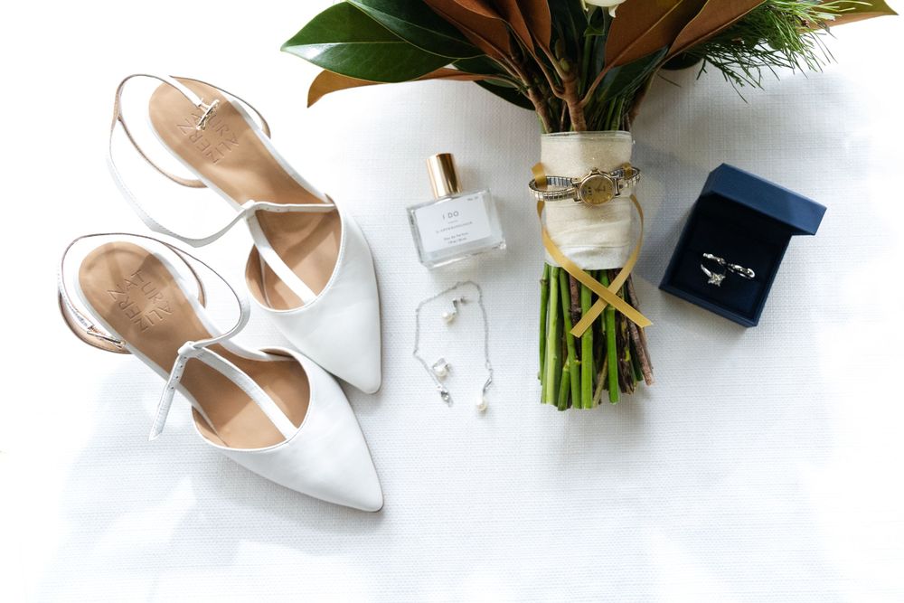 Wedding accessories including white heels, perfume bottle, flower bouquet and navy ring box arranged on white background.
