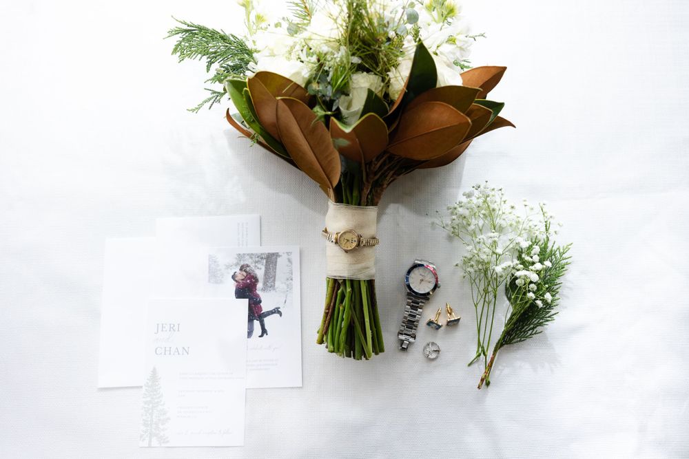 Wedding details flatlay with bouquet featuring magnolia leaves, rings, and floral arrangements on white surface.