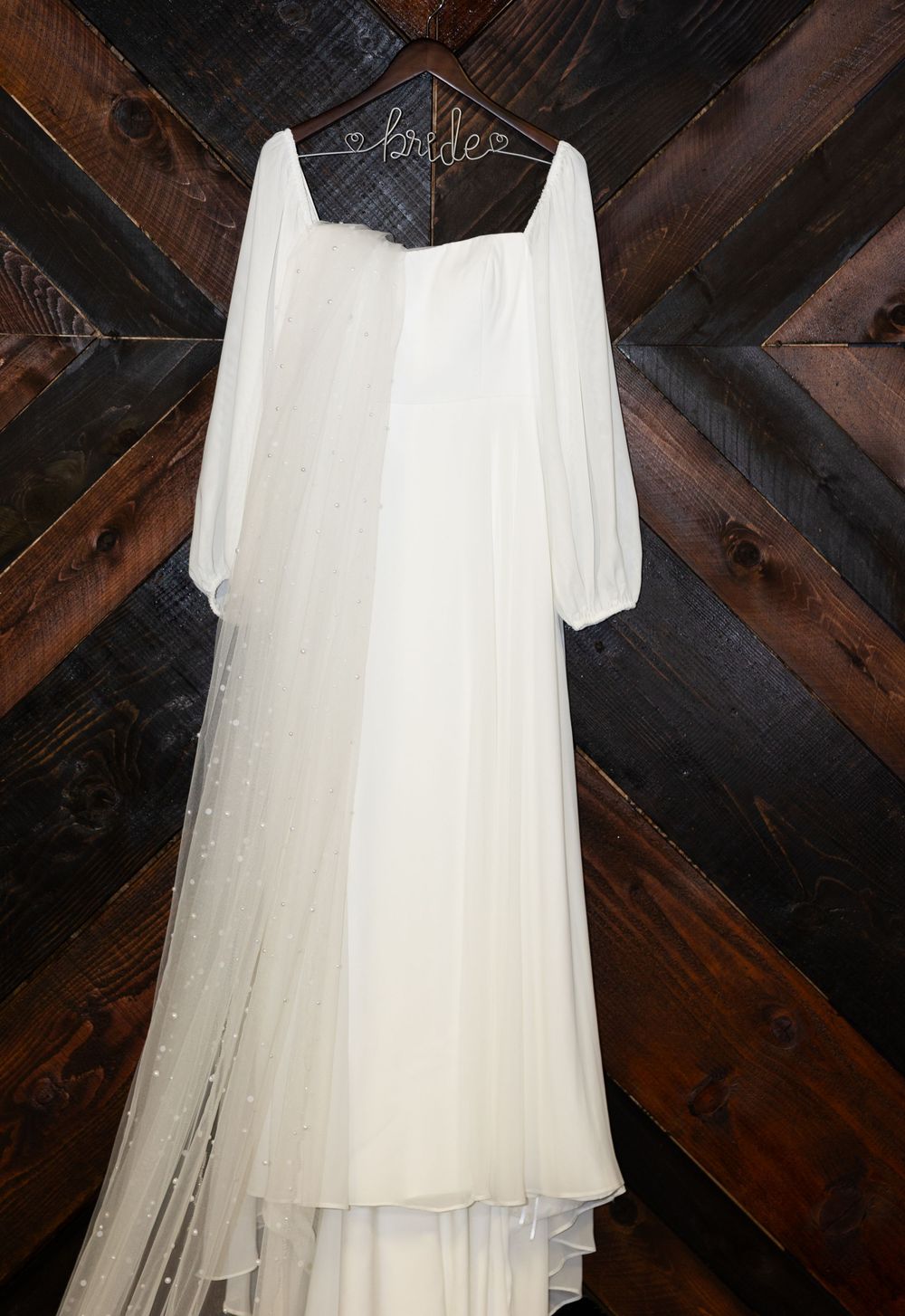An elegant white wedding dress with long sleeves hangs on a wooden wall.