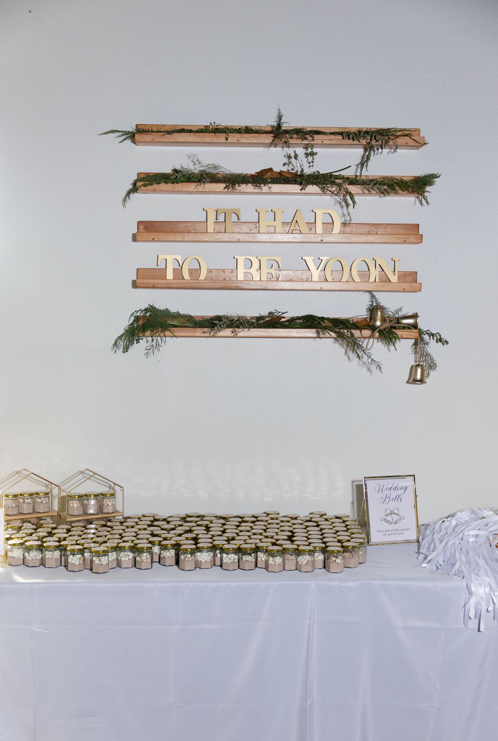 Rustic wooden sign display reading 'IN HEAD TO BE YOON' with evergreen branches and rows of favors below.