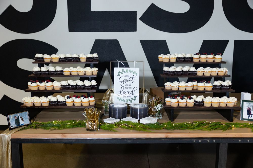 Wedding dessert display table with rows of mini cupcakes arranged on wooden stands against a black wall.