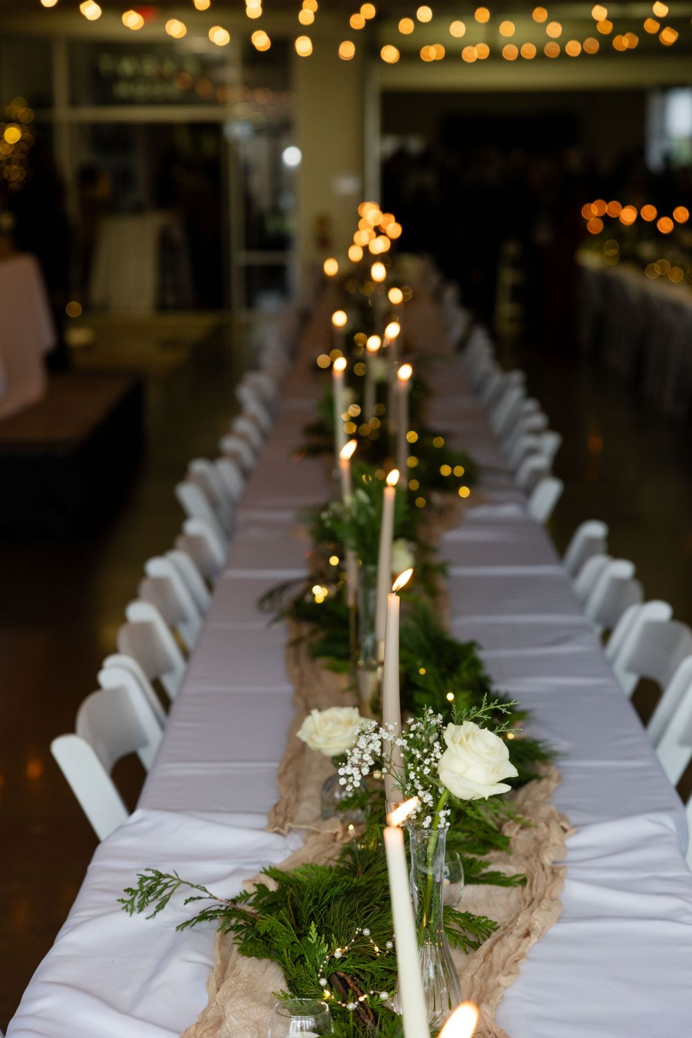 Long reception table set with white linens, greenery runners, and taper candles under string lights.