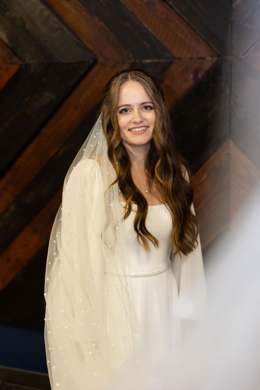 A bride in a simple white wedding gown and veil against a wooden wall backdrop.