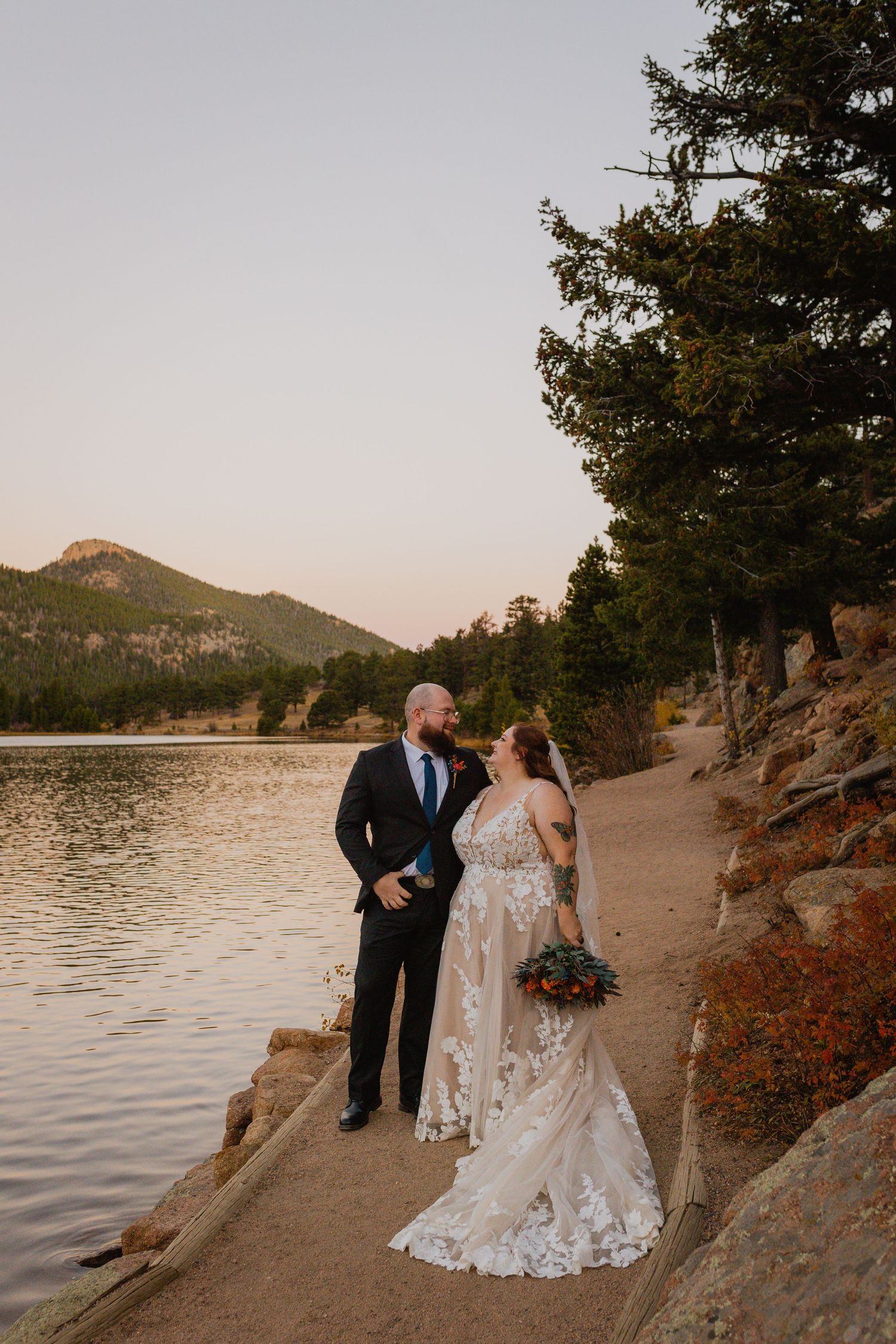 A couple shares a romantic moment at sunset by a mountain lake, standing on a rocky shoreline path.