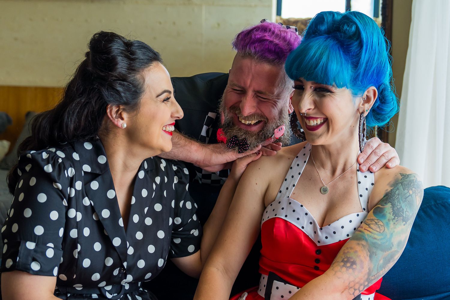 Three friends laugh together while sitting on a blue couch, one wearing polka dots and another in a red top.