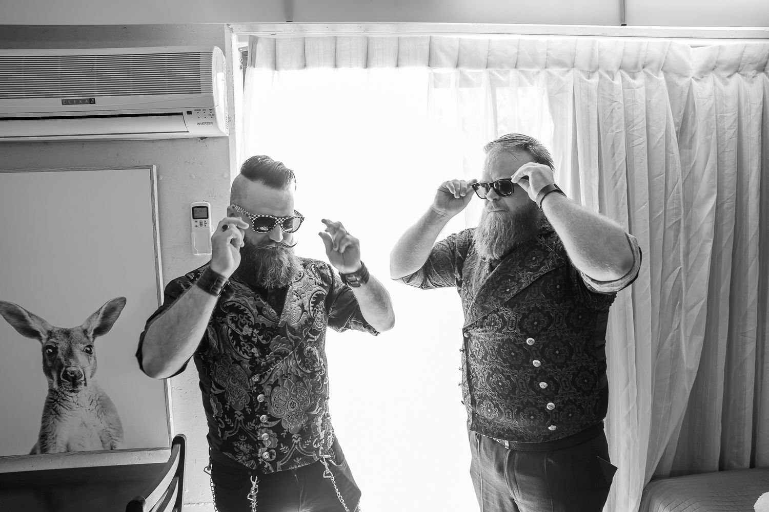 A black and white image showing two people in floral vests posing playfully with a kangaroo in a room.