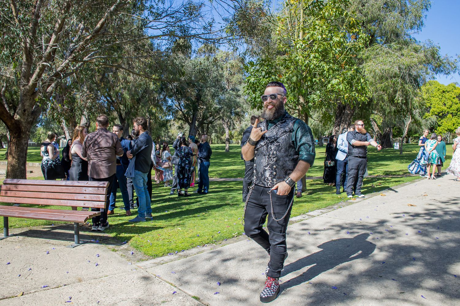 Group of people gathered in a sunny park with walking path and benches while someone speaks into a microphone.
