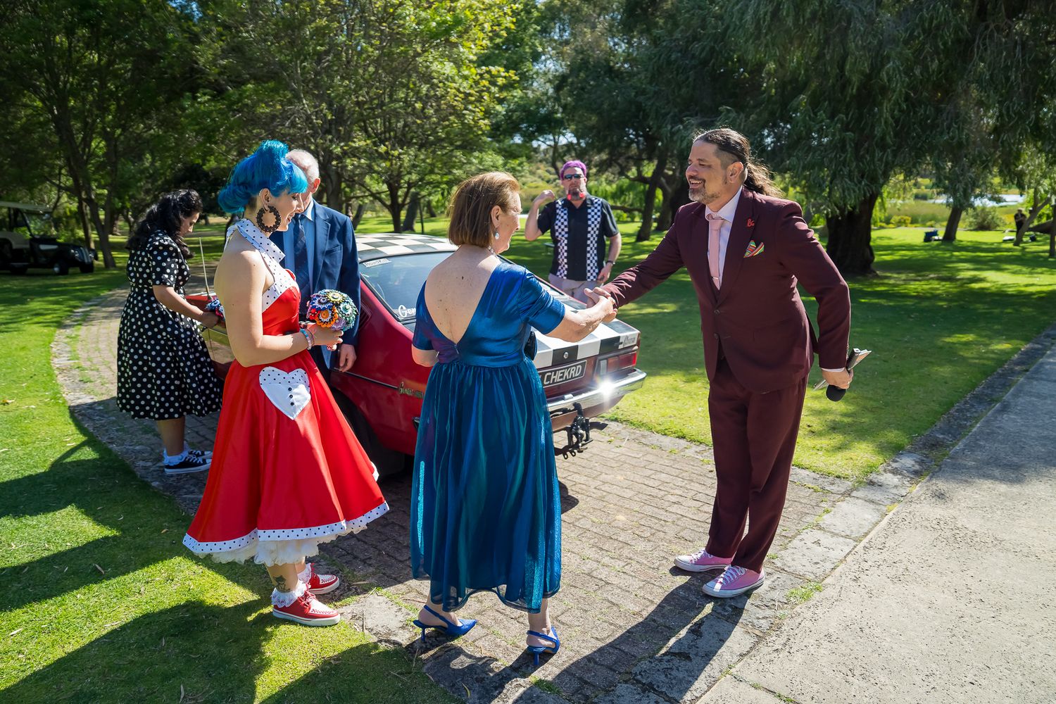 A group of people in vintage dresses and formal wear gather on a sunny path in a lush green park.