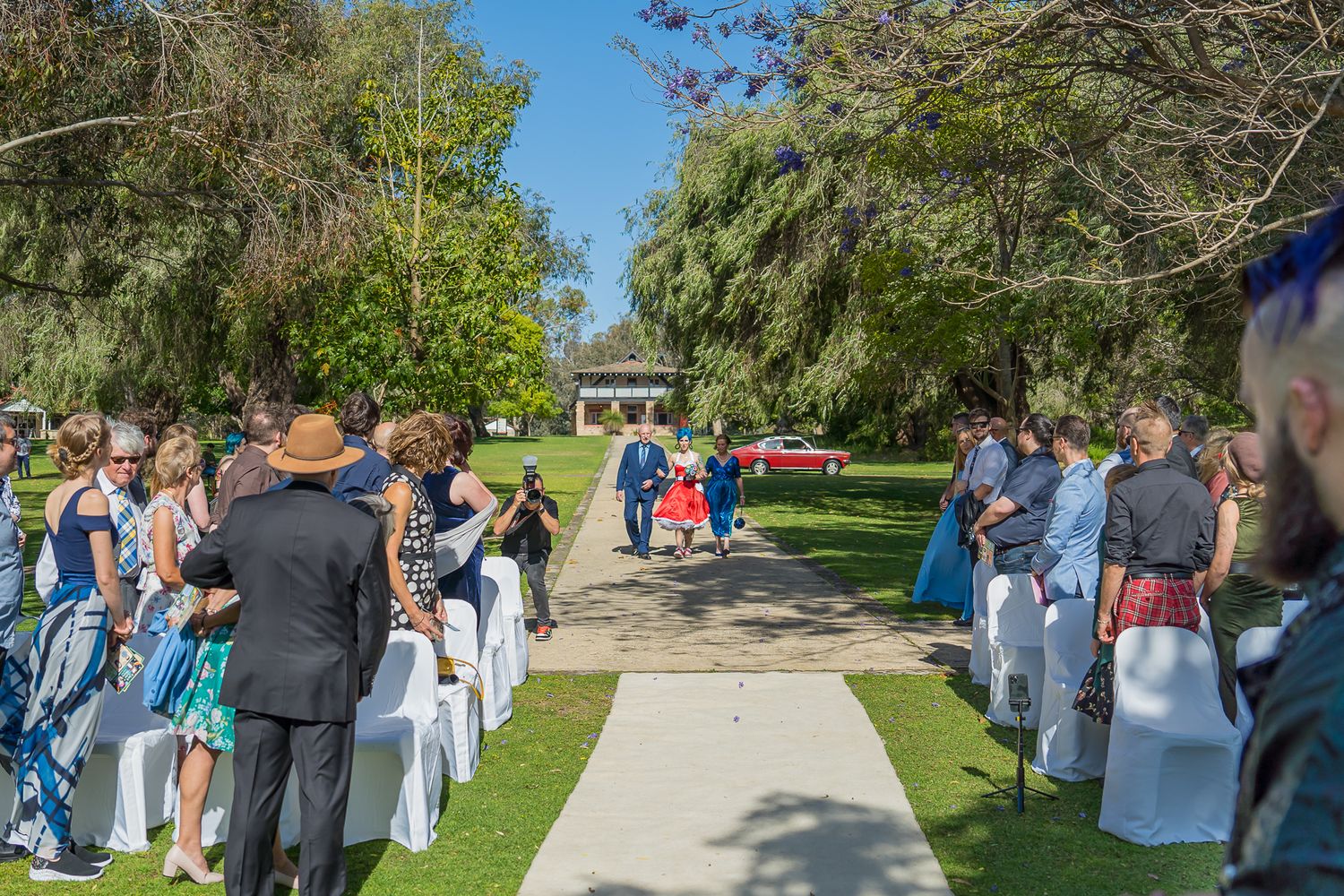 Guests gather along a walkway at an outdoor wedding ceremony under blue skies and trees.