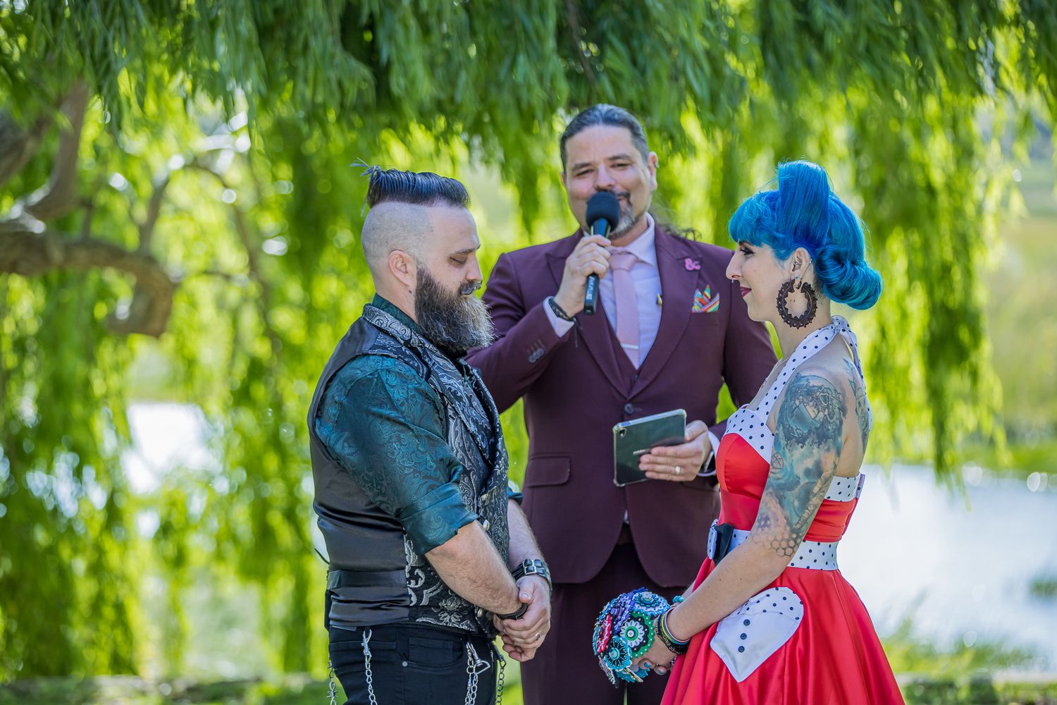 A wedding ceremony takes place under a green willow tree with burgundy and red attire worn by the participants.