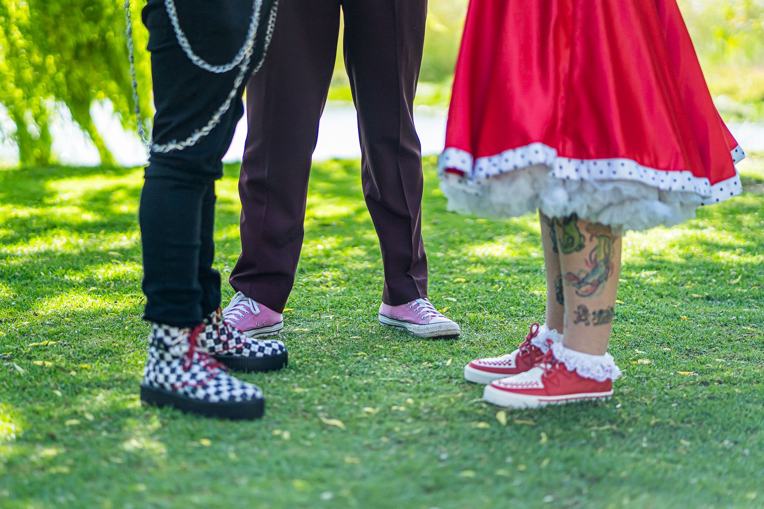 Two pairs of feet wearing Converse sneakers - one checkered and one red - standing on grass with a red dress visible.