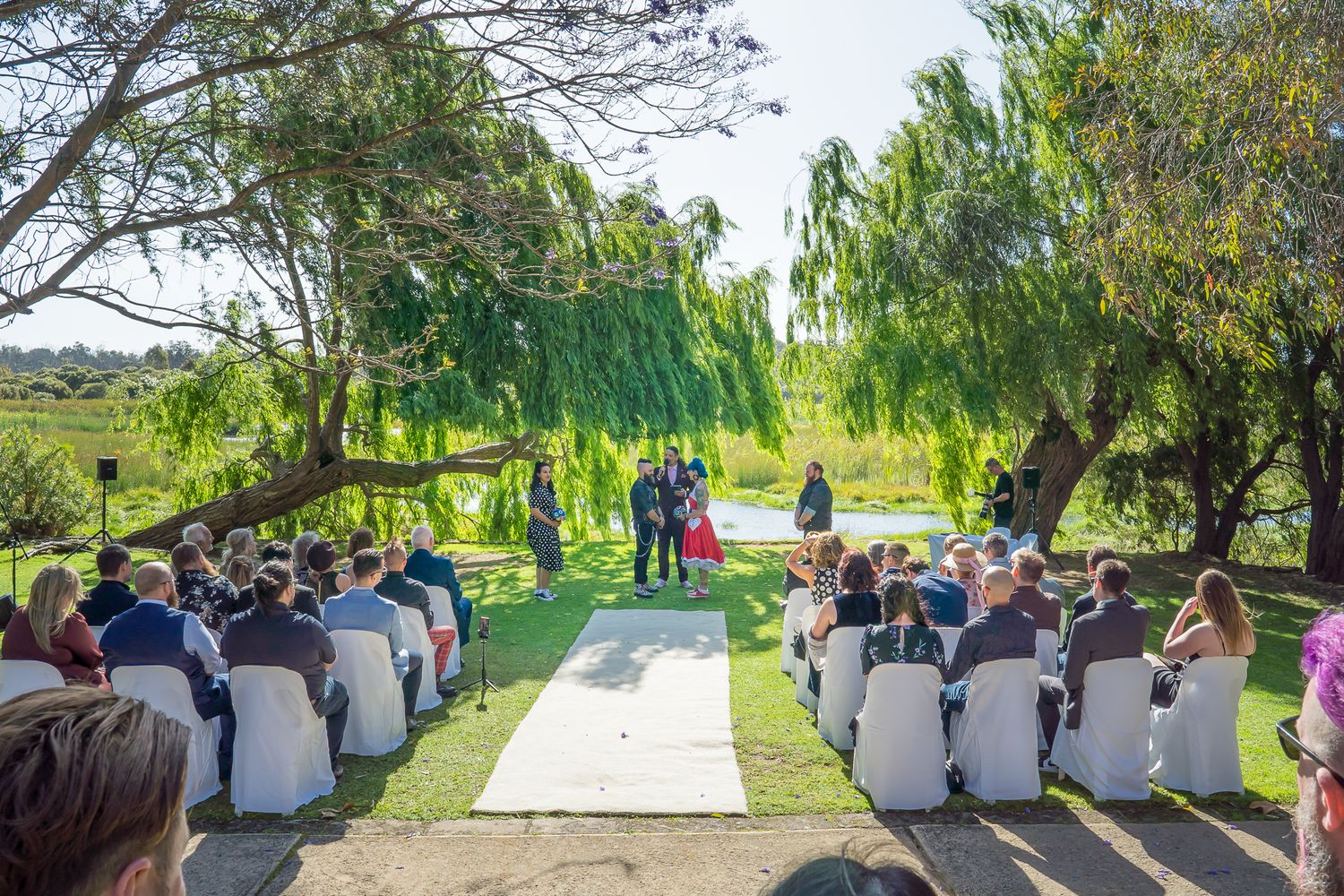 Outdoor garden wedding ceremony with white chairs and willow trees creating a beautiful natural setting.